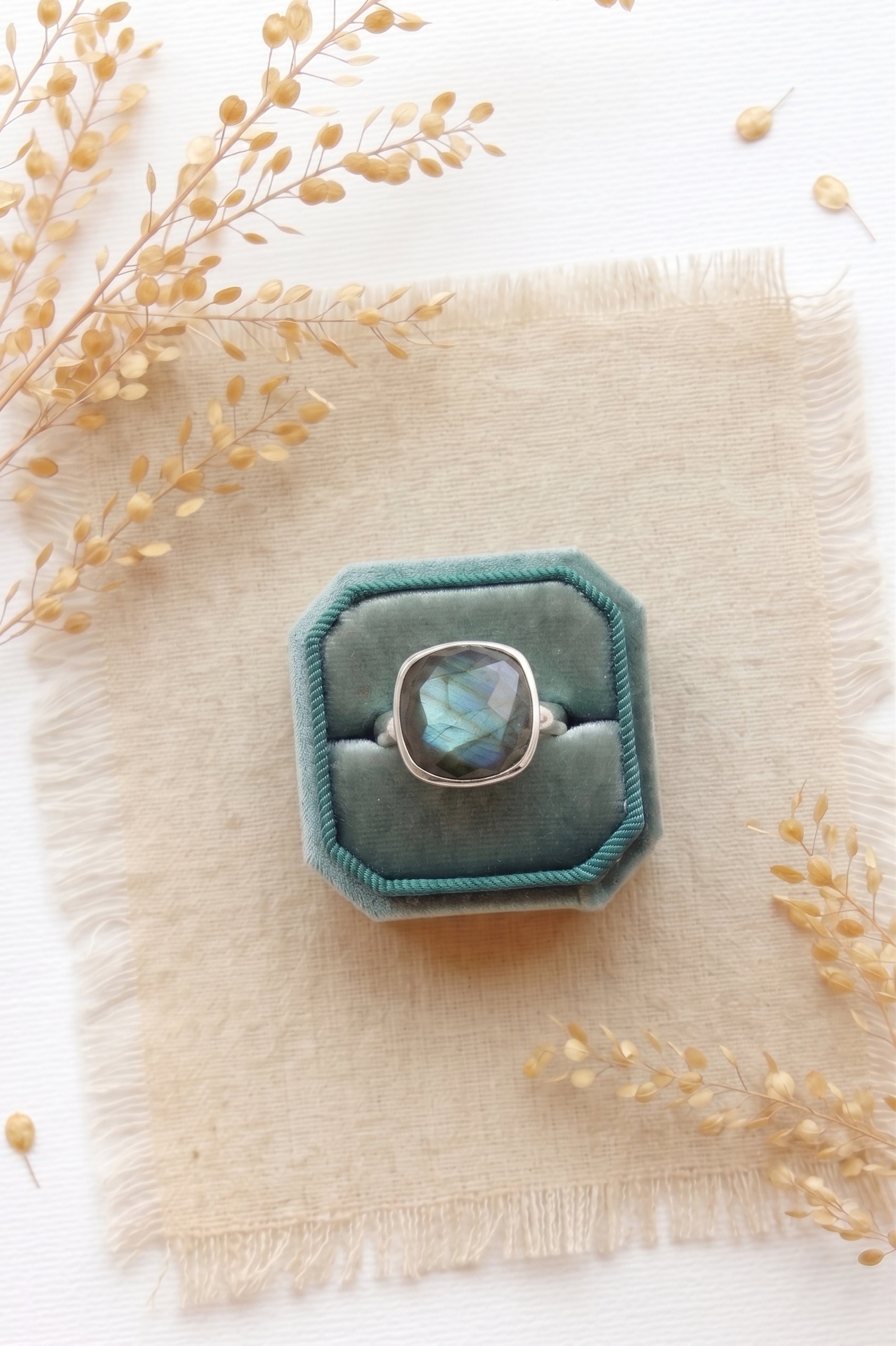 Portrait of a blonde woman wearing a deep teal velvet dress standing in a sunlit field of golden grass. She holds one hand up to her chin, elegantly showcasing a prominent, flashing blue cushion-cut labradorite silver cocktail ring on her middle finger.