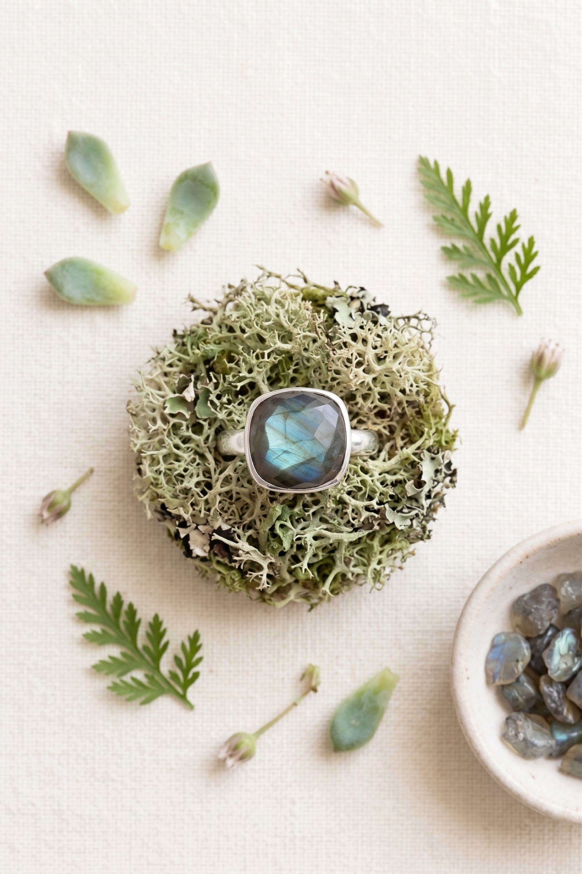 Flat lay of a checkered-facet cushion labradorite silver ring centered on a round clump of green reindeer moss. The background features a white linen texture styled with scattered green succulent leaves, small flower buds, and a ceramic bowl filled with raw labradorite chips.
