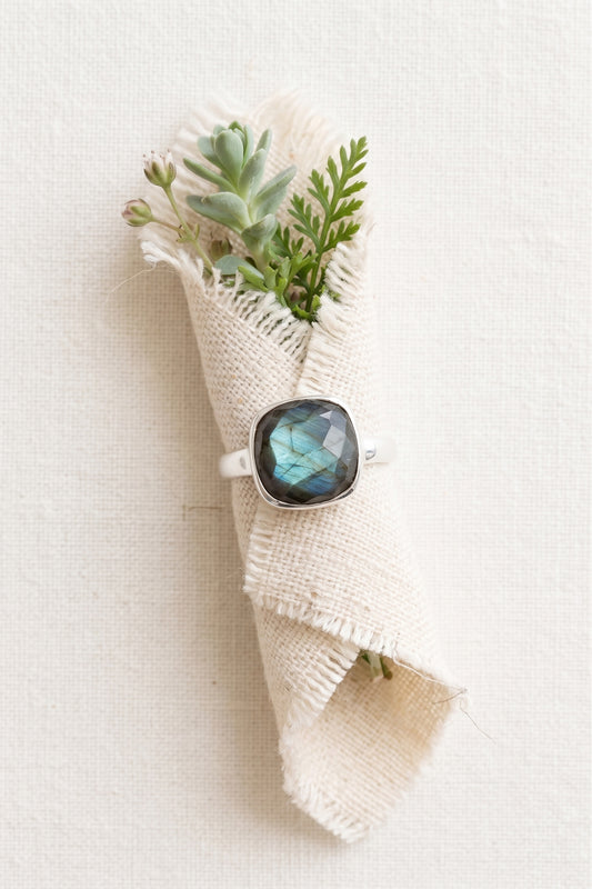 A sterling silver labradorite ring with a prominent teal-blue labradorescence flash, wrapped around rolled linen fabric filled with a small bouquet of green succulents and delicate sprigs, shot from directly above against a bright fabric background.