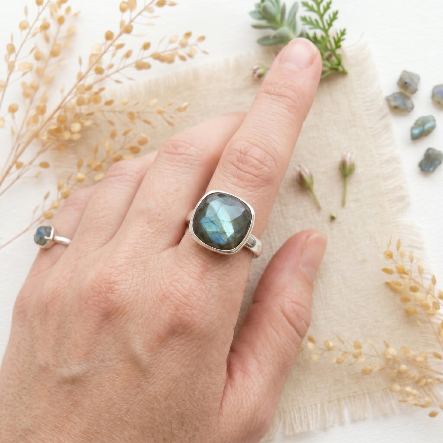 A person wearing a large, cushion-cut labradorite ring with a vibrant blue-green flash on their index finger, set in a smooth sterling silver bezel. The hand rests on a rustic linen cloth surrounded by dried botanicals, small raw labradorite crystals, and a delicate silver pinky ring. 
