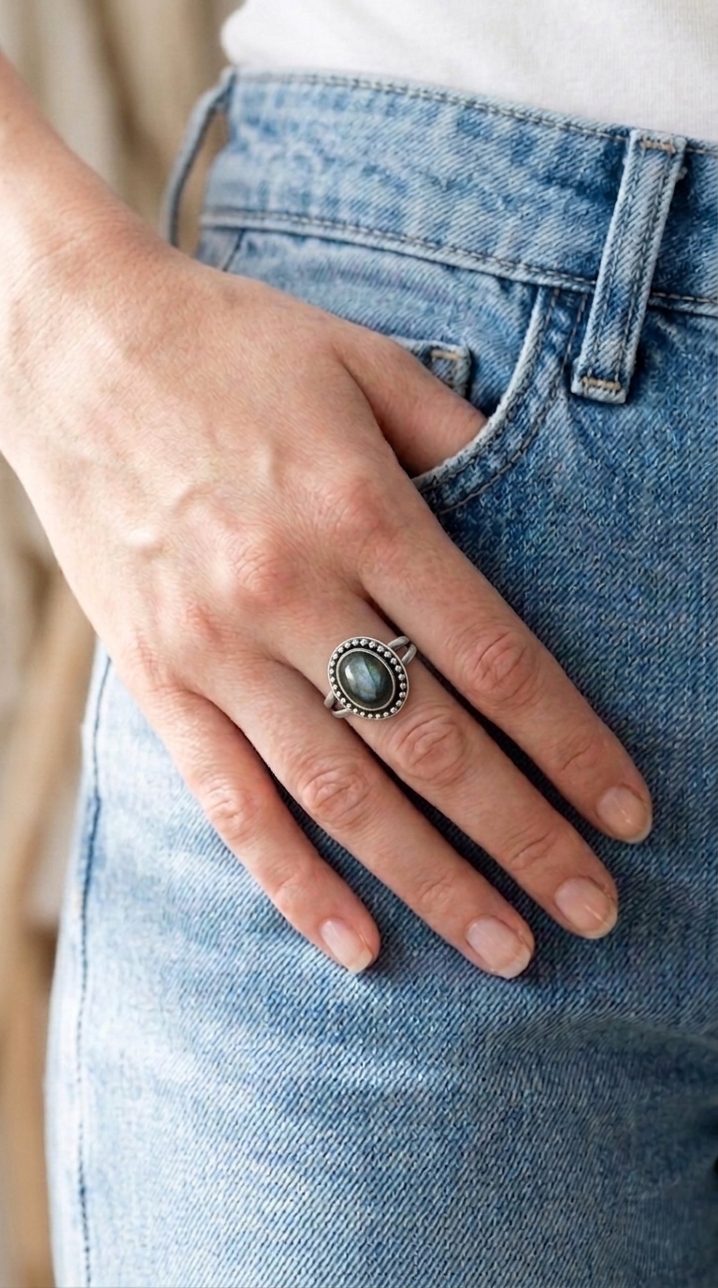 A sterling silver ring with a large, oval, iridescent labradorite gemstone. The gemstone is surrounded by a beaded border. Worn by a woman with her hand tucked partially in her pocket.