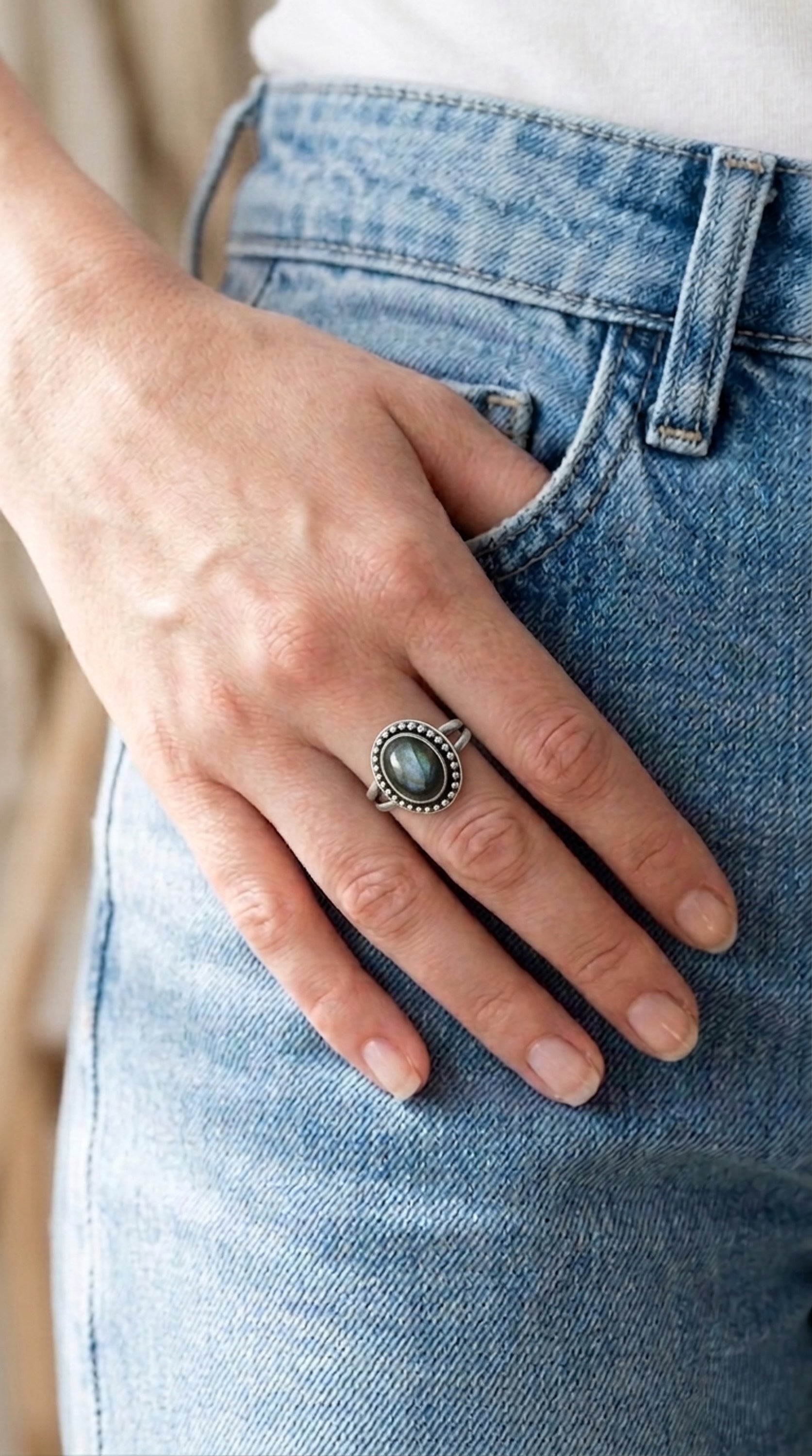 A sterling silver ring with a large, oval, iridescent labradorite gemstone. The gemstone is surrounded by a beaded border. Worn by a woman with her hand tucked partially in her pocket.