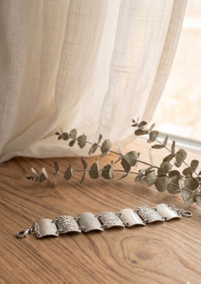 A Sterling Silver bracelet on a wooden surface with a light curtain background. The bracelet features wide, curved segments, each adorned with detailed floral and geometric engravings. The segments are connected by small metal loops, and a clasp is visible on the left side. 

