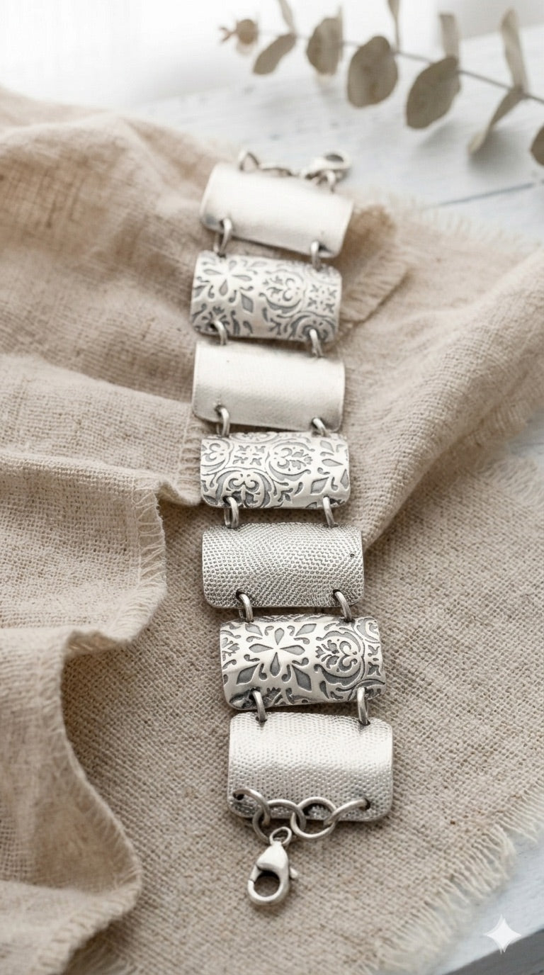 A Sterling Silver bracelet on a wooden surface with a light curtain background. The bracelet features 7, 1.25 inch x 3/4 inch wide, curved segments, each adorned with detailed floral and geometric engravings. The segments are connected by small metal loops, and a clasp is visible on the left side. 
