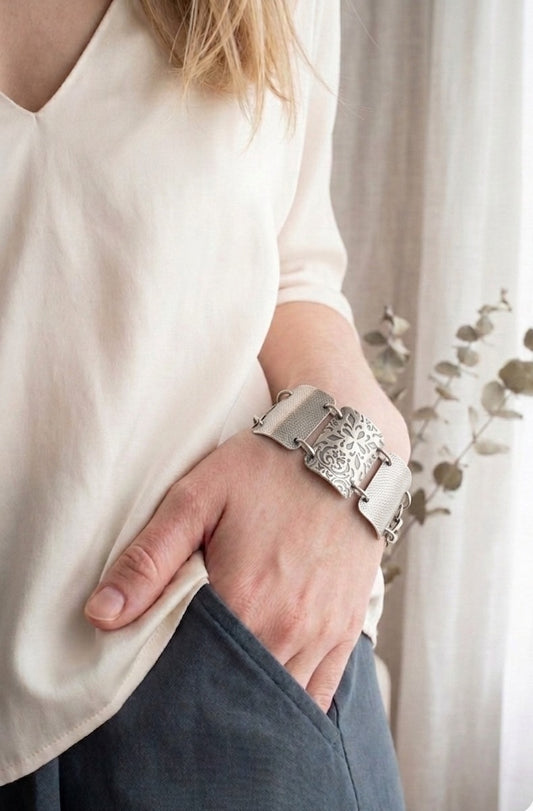 Close-up of sterling silver rectangle links showing oxidized and brushed texture detail. The bracelet features 7, 1.25 inch x 3/4 inch wide, curved segments, each adorned with detailed floral and geometric engravings. The segments are connected by small metal loops, and a lobster clasp.
