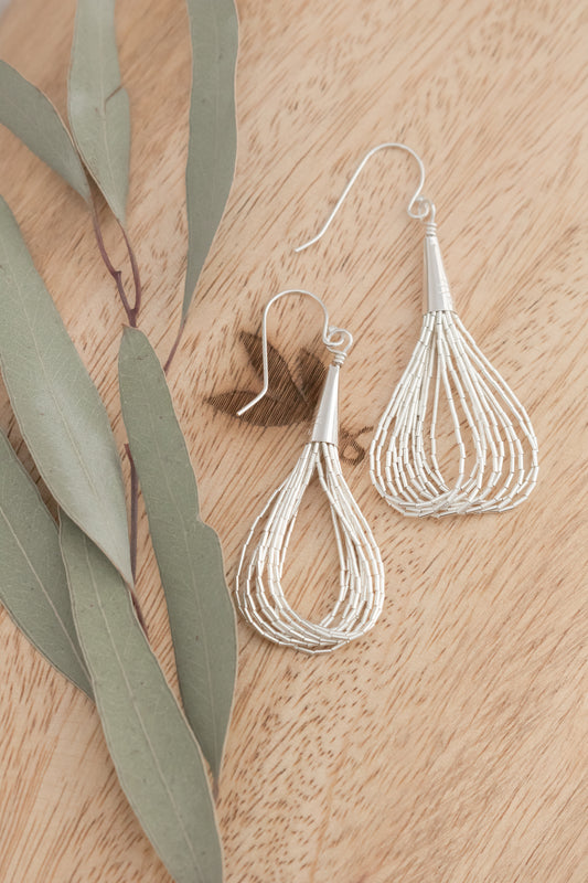 Angled shot of two silver cascading earrings placed on a wooden surface with eucalyptus leaves, capturing their movement and shimmering quality.

