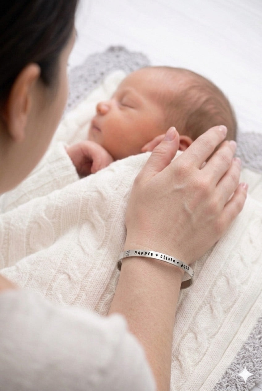 A personalized sterling silver name cuff bracelet worn on a mother's wrist as she rests her hand on a sleeping newborn. The slim, polished cuff features the names "Harper," "Olivia," and "Jack" in a dainty, hand-stamped font with small heart accents between the names and fern details at both ends.