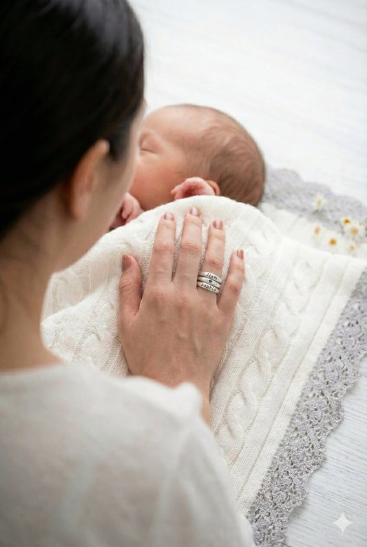 Woman wearing custom sterling silver name rings holding a baby wrapped in a white blanket on a light surface