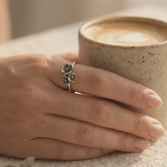 A hand wearing a handcrafted sterling silver ring with a cluster of three small textured flowers, holding a ceramic coffee cup as warm sunlight illuminates the ring's design.
