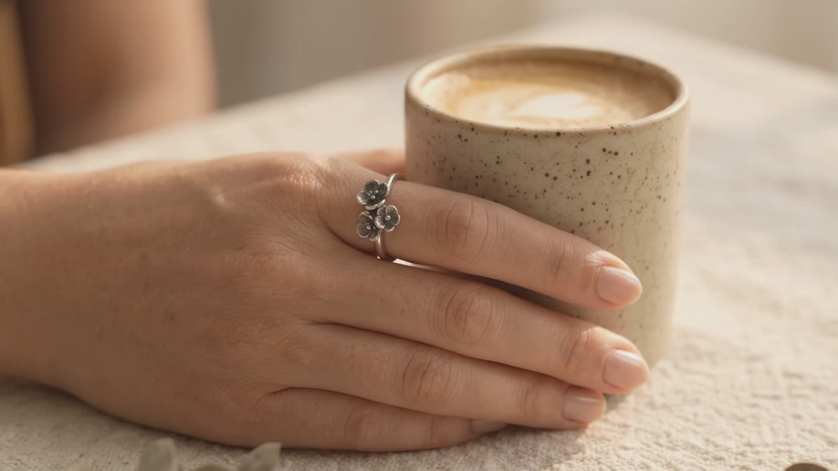 A hand wearing a handcrafted sterling silver ring with a cluster of three small textured flowers, holding a ceramic coffee cup as warm sunlight illuminates the ring's design.