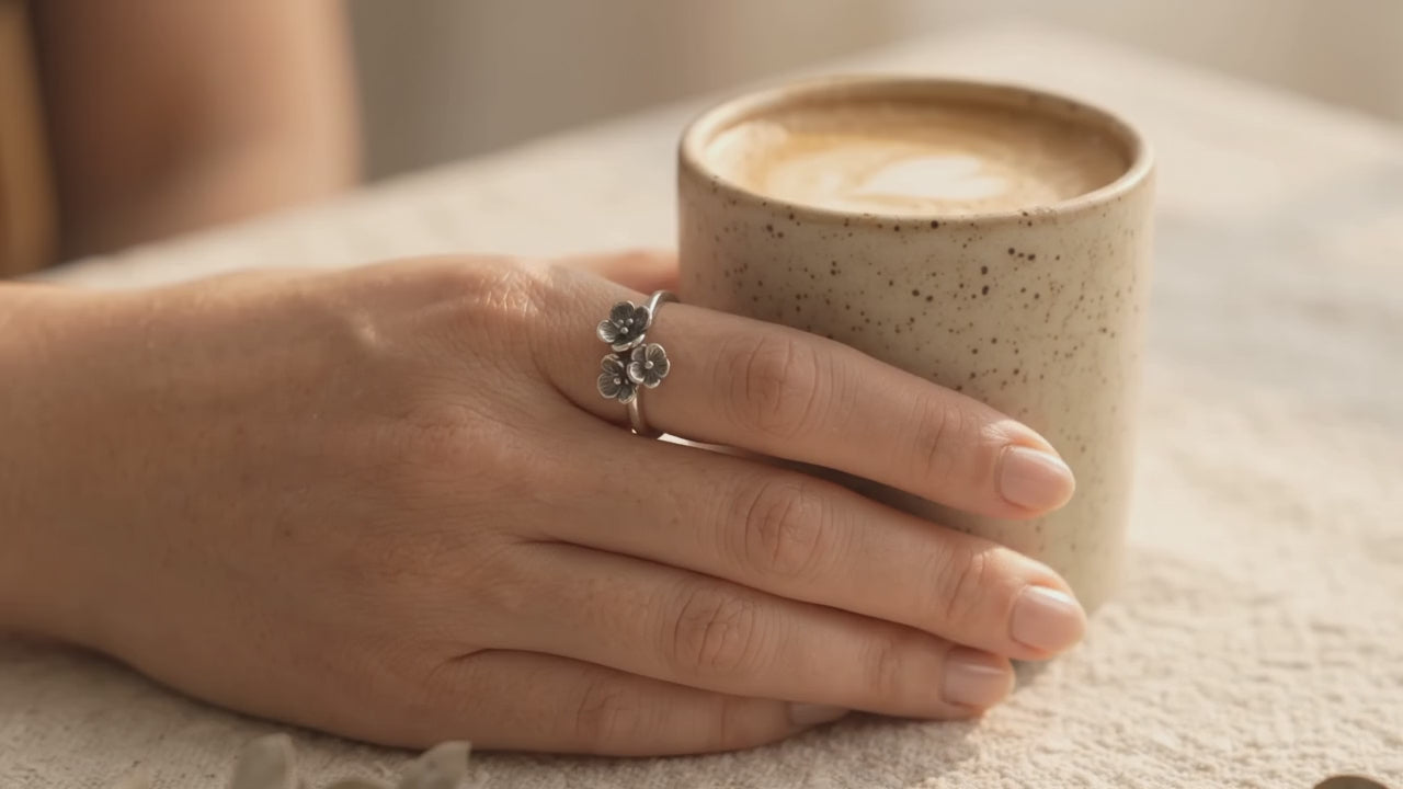 A hand wearing a handcrafted sterling silver ring with a cluster of three small textured flowers, holding a ceramic coffee cup as warm sunlight illuminates the ring's design.