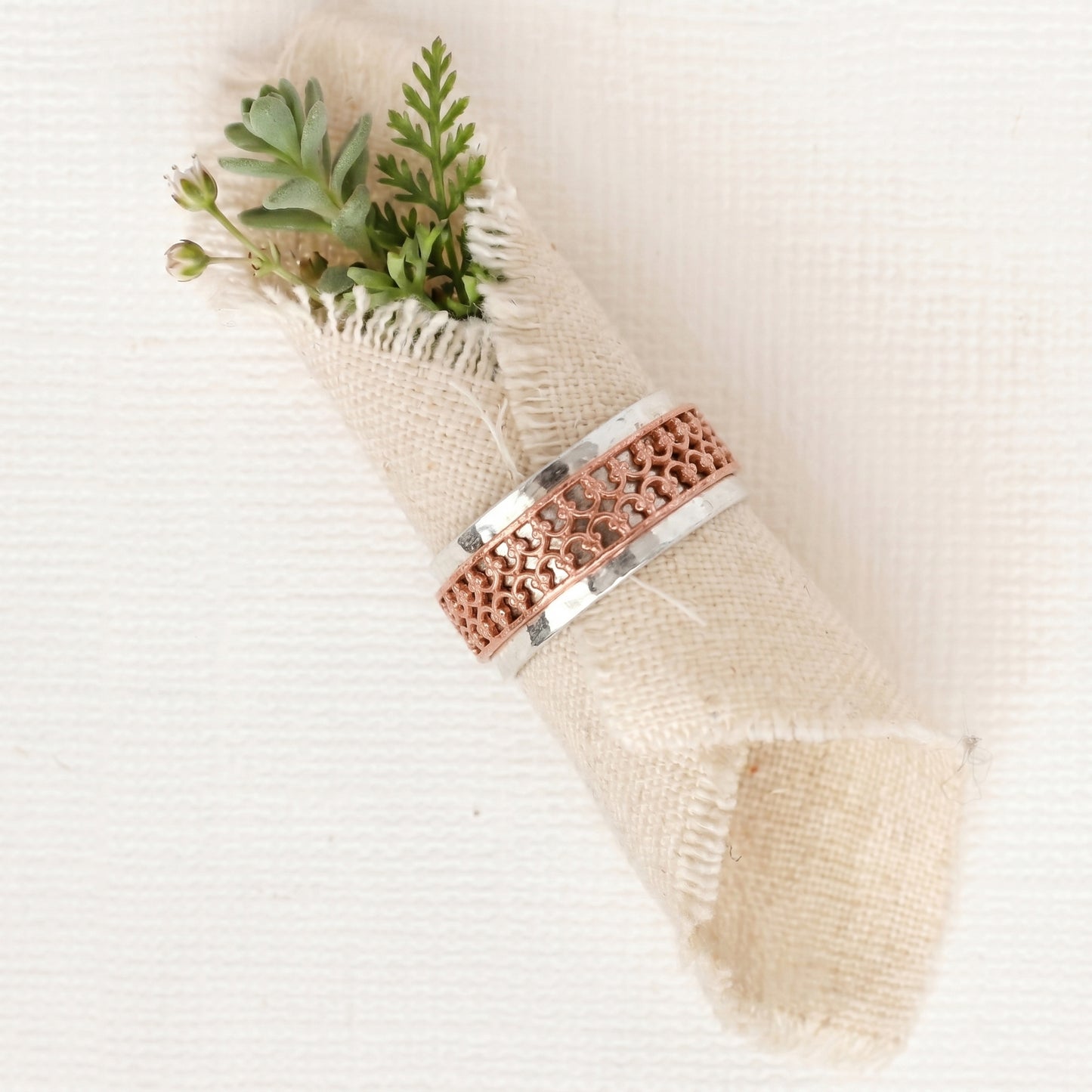 Two-tone ring with a sterling silver hammered band and an outer double crown wire copper band the spins freely on a light fabric background with green leaves.