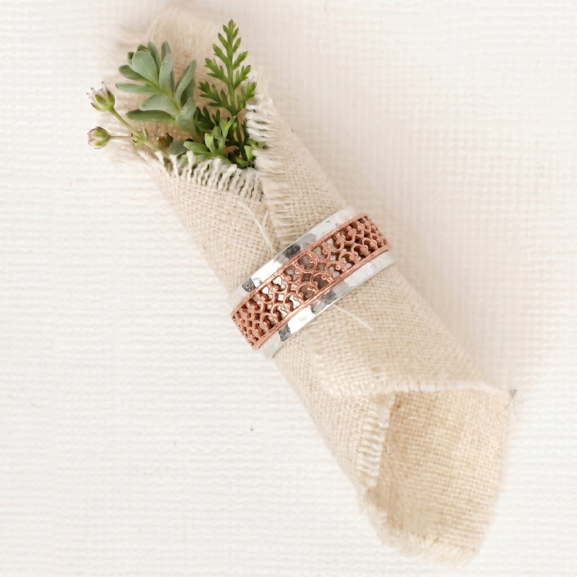 Two-tone ring with a sterling silver hammered band and an outer double crown wire copper band the spins freely on a light fabric background with green leaves.