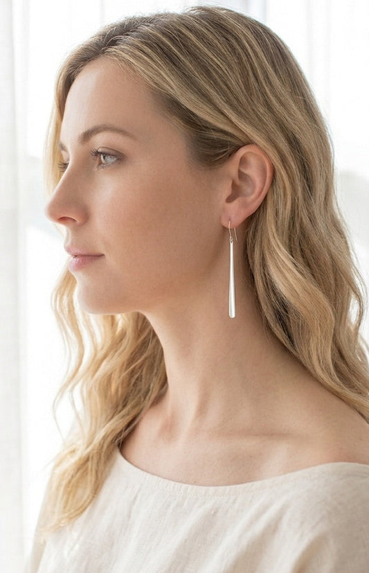 The model is wearing a long, minimalist polished sterling silver long drop earrings. The earring is a clean, elongated bar that tapers from 1.5mm at the top to 3mm at the base and is approximately 2.25 inches in length.