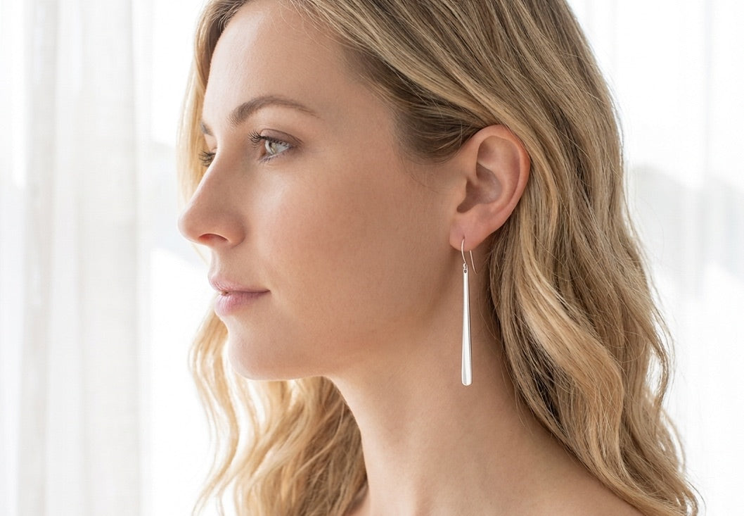 The model is wearing a long, minimalist polished sterling silver dangle earring. The earring is a clean, elongated bar that tapers from 1.5mm at the top to 3mm at the base and is approximately 2.25 inches in length.