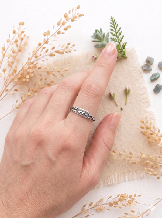 A hand wearing a sterling silver floral band ring on the index finger, set against a soft flat lay background with dried botanical sprigs, small pink flower buds, and raw gemstones on a frayed linen coaster.
