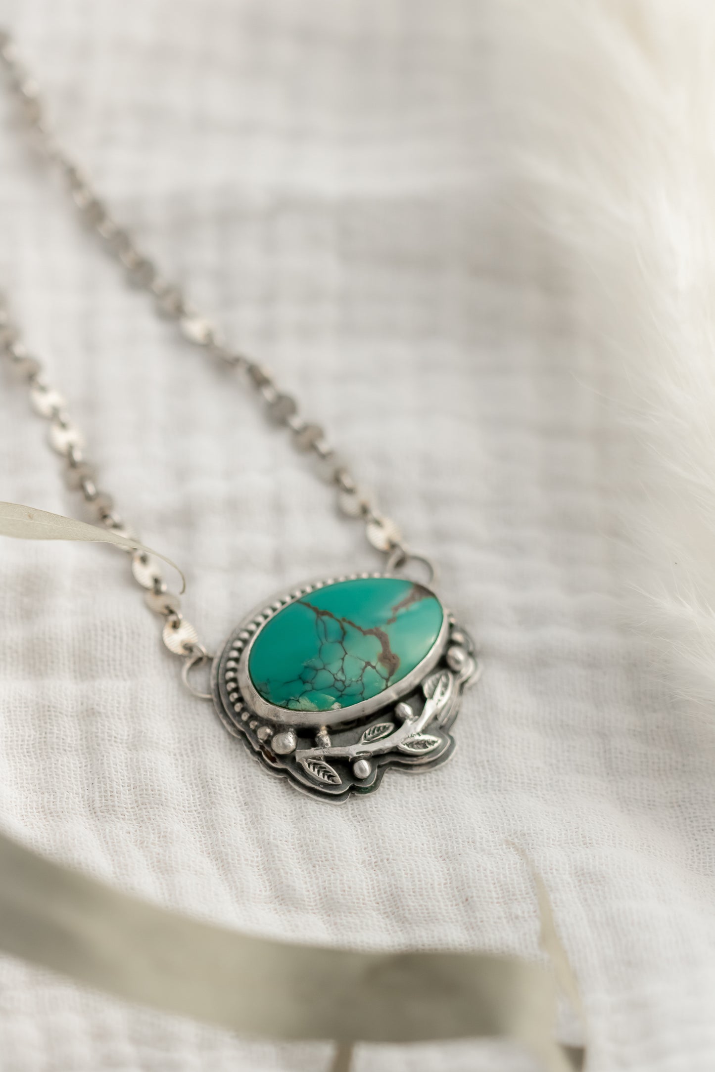 Handcrafted turquoise pendant necklace in sterling silver with leaf design on a white fabric background 
