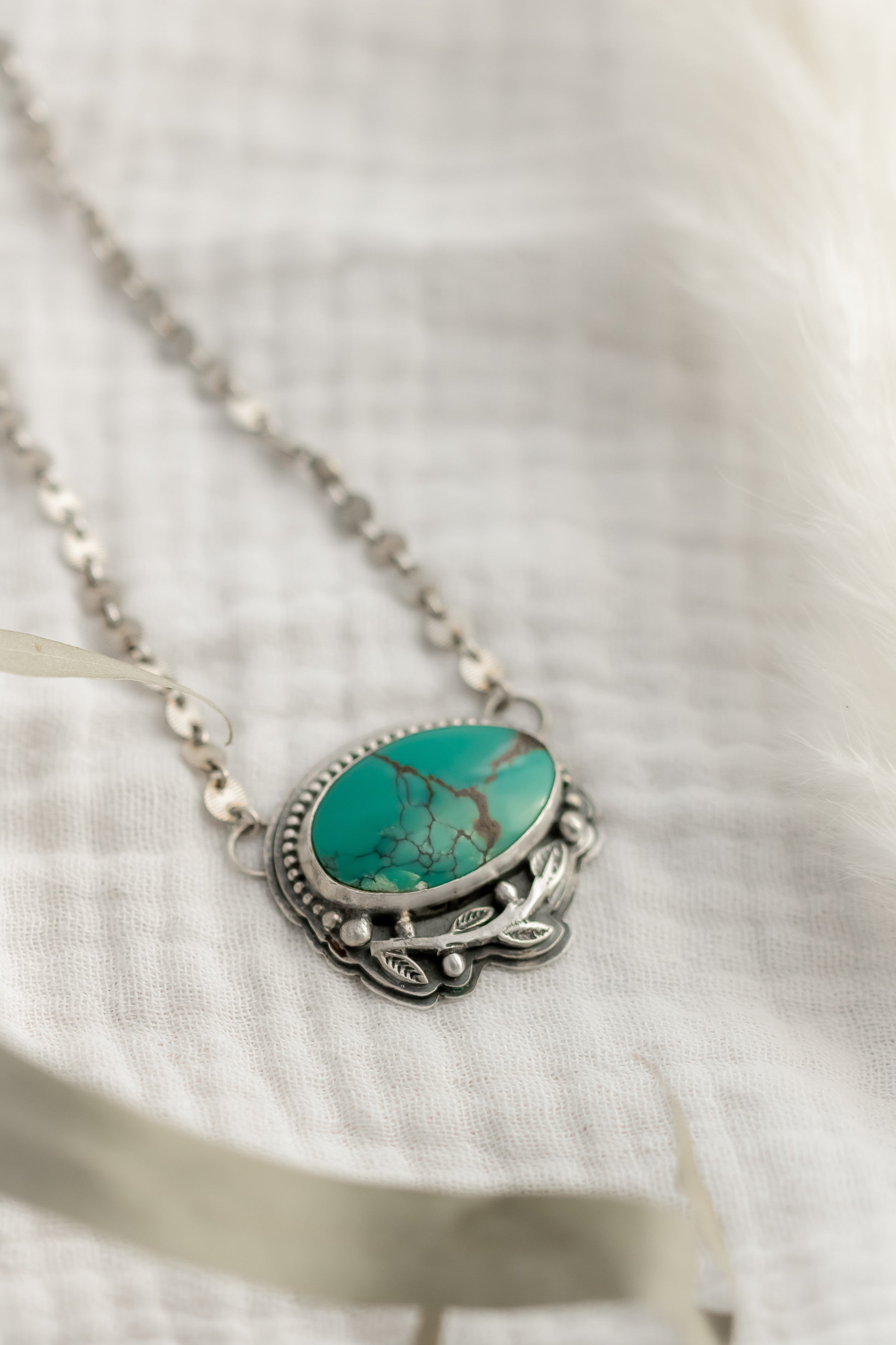 Handcrafted turquoise pendant necklace in sterling silver with leaf design on a white fabric background 
