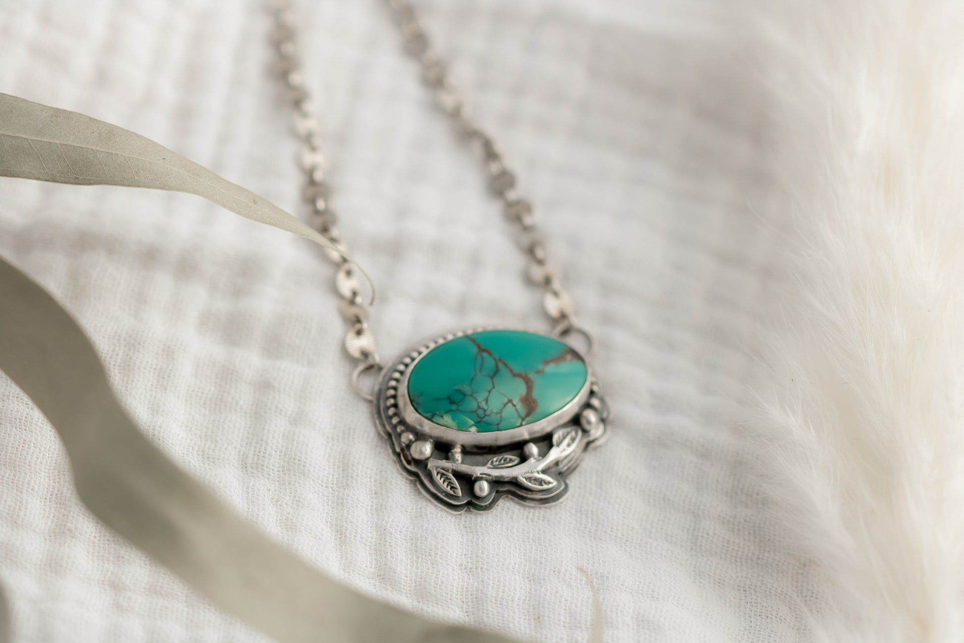 Handcrafted turquoise pendant necklace in sterling silver with leaf design on a white fabric background 
