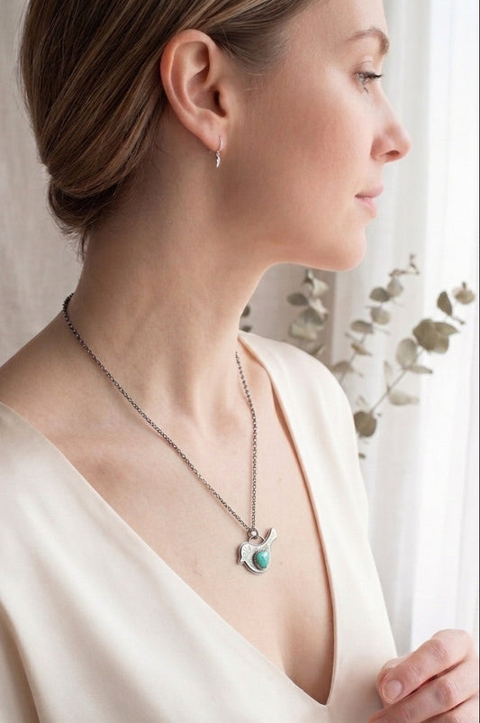 Woman wearing a sterling silver bird shaped pendant necklace with floral design and turquoise wing against a neutral background.