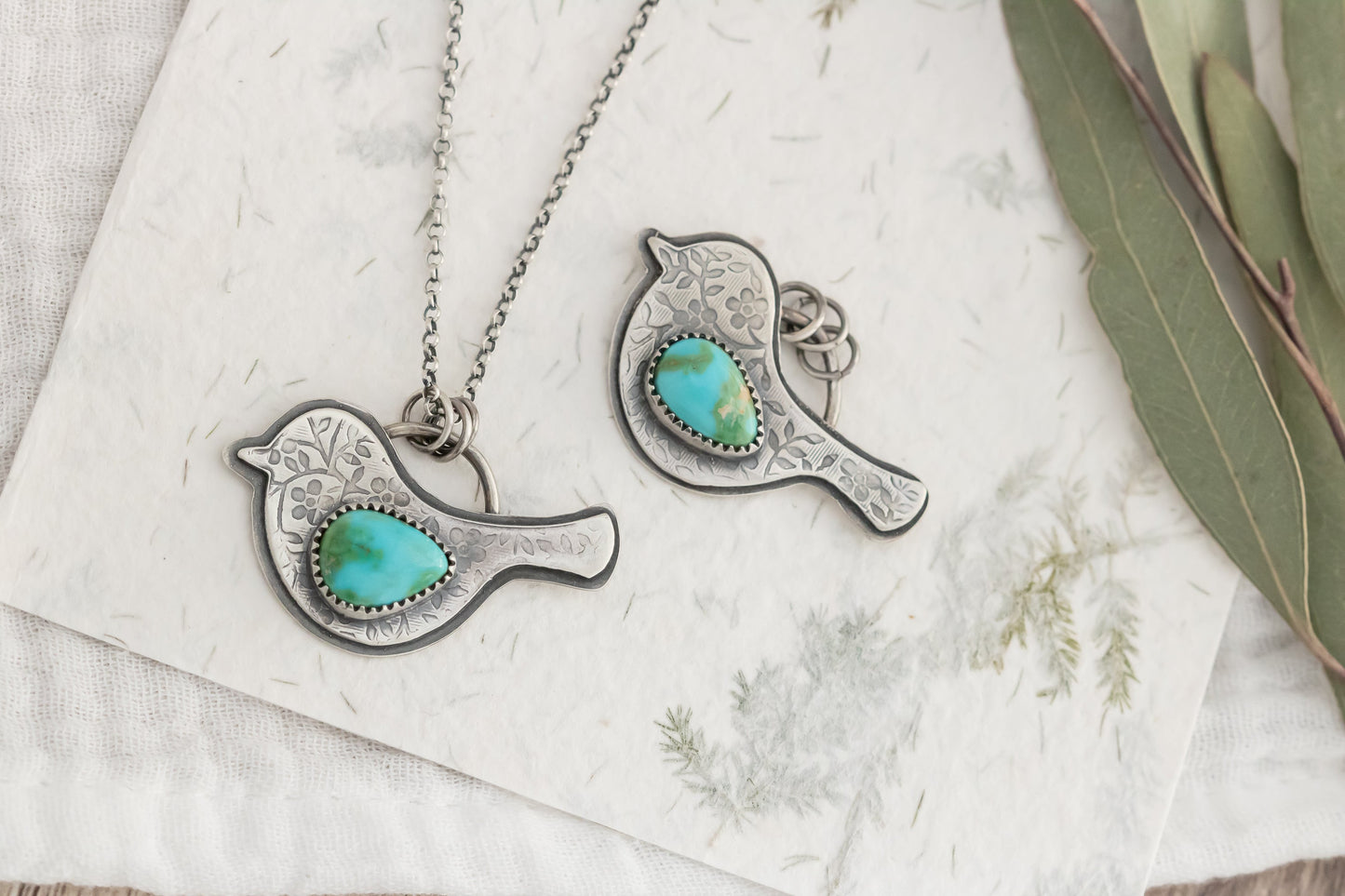 Turquoise stone bird pendants on silver chains on a textured white surface with greenery.