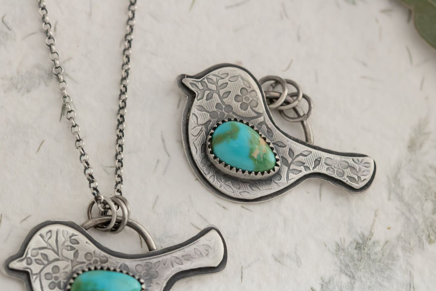 Silver bird-shaped pendant with floral texture and turquoise stone on a chain against a light background
