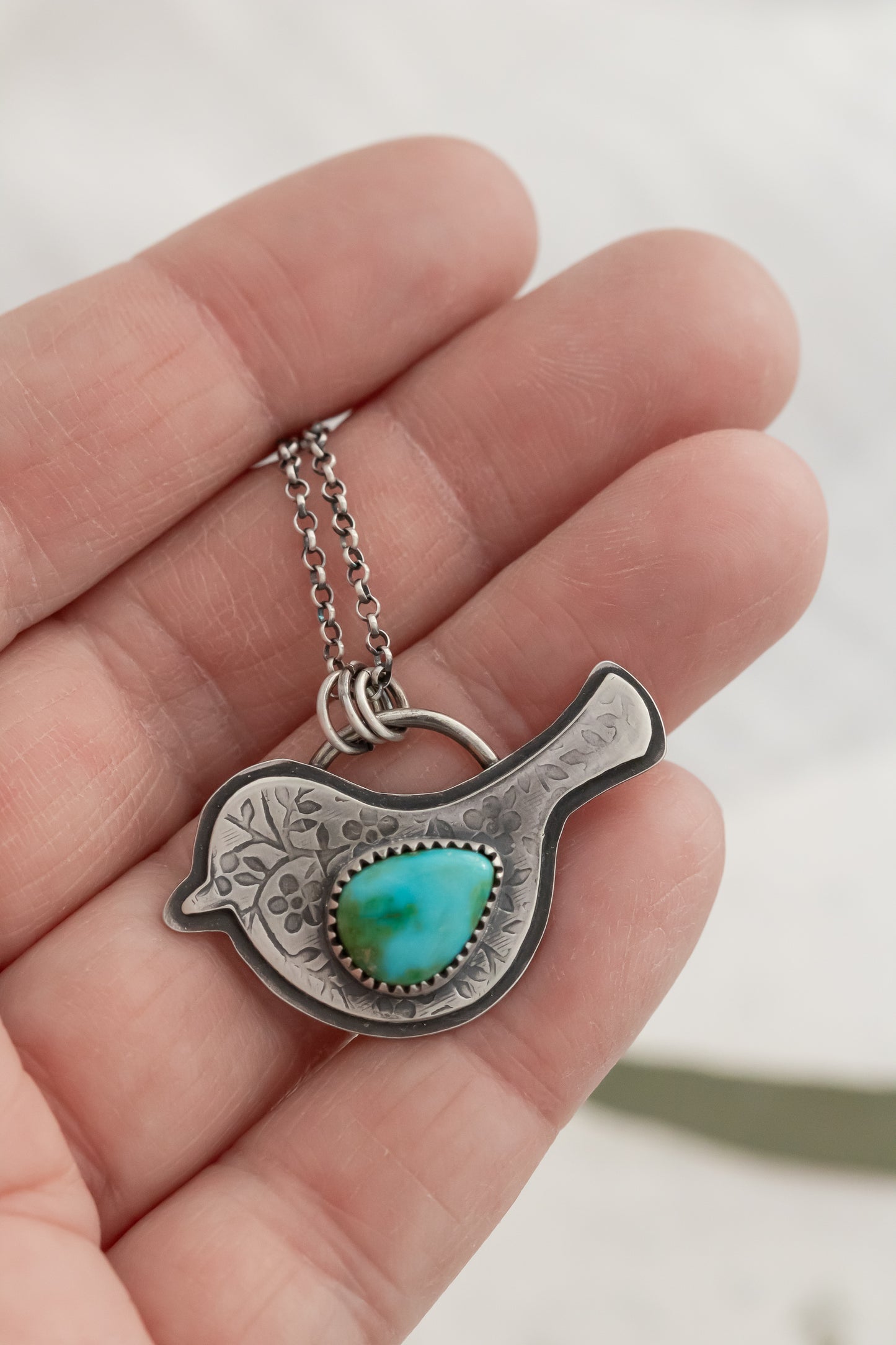 Hand gently holding sterling silver bird necklace with vibrant turquoise stone against neutral backdrop  
