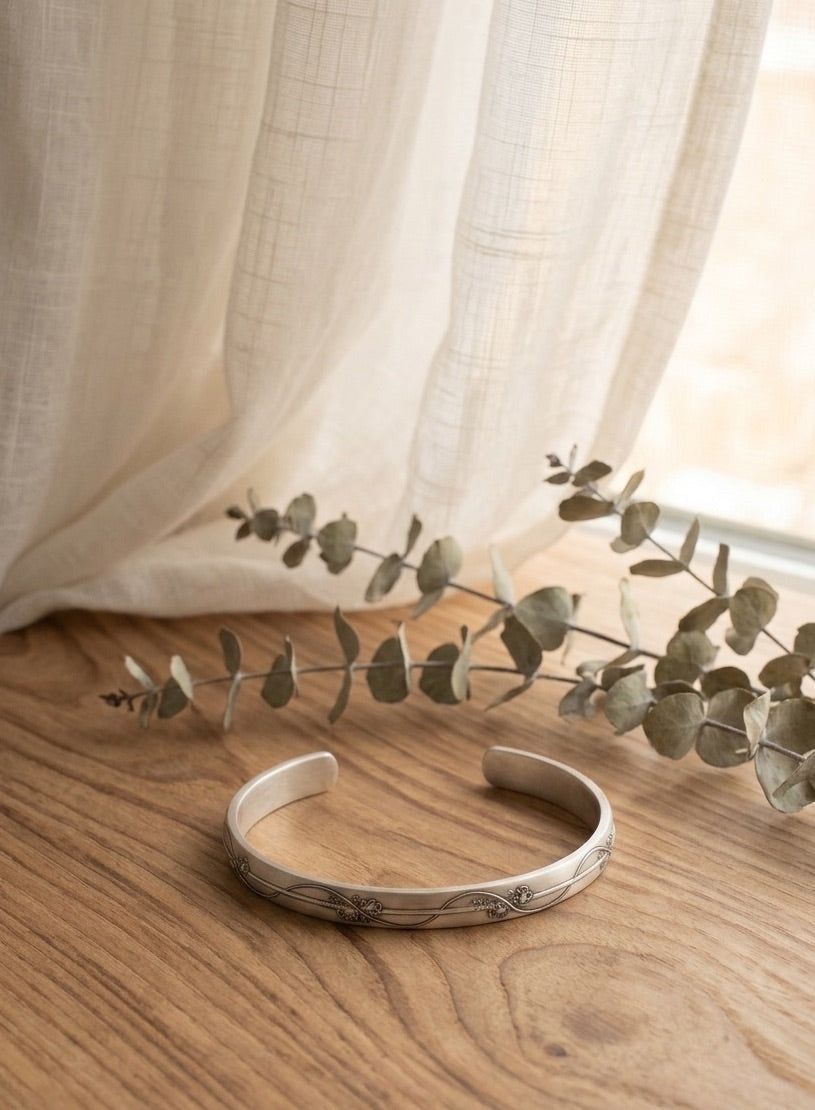 Sterling silver bracelet with a floral vine design on a wooden surface with eucalyptus leaves and sheer curtains in the background