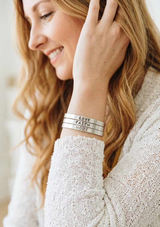Woman wearing silver bracelets with 'LOVE', 'FAITH', and 'HOPE' inscriptions on a blurred background.