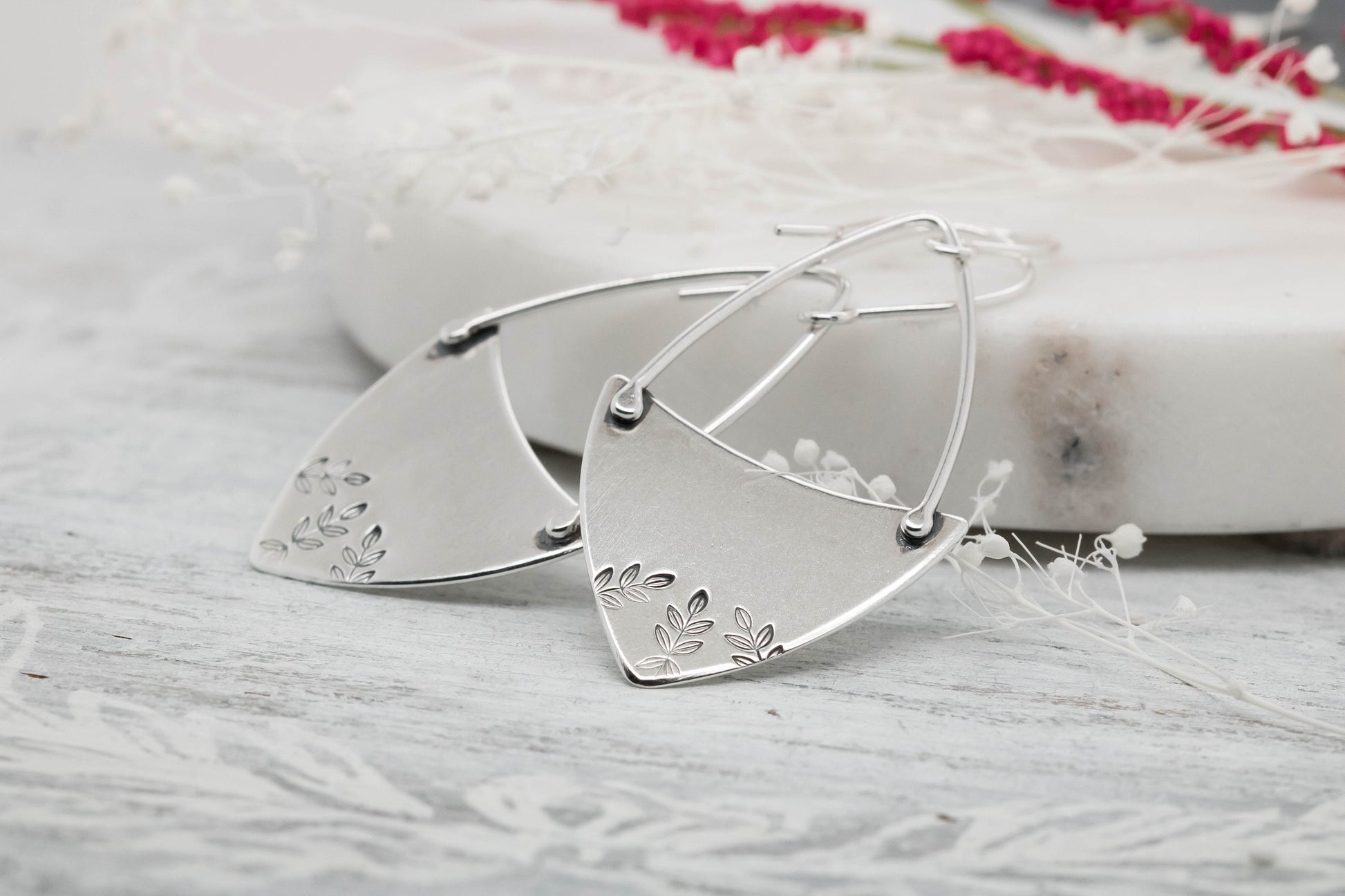 A pair of sterling silver hoop earrings with a floral fern design, displayed against a natural backdrop.