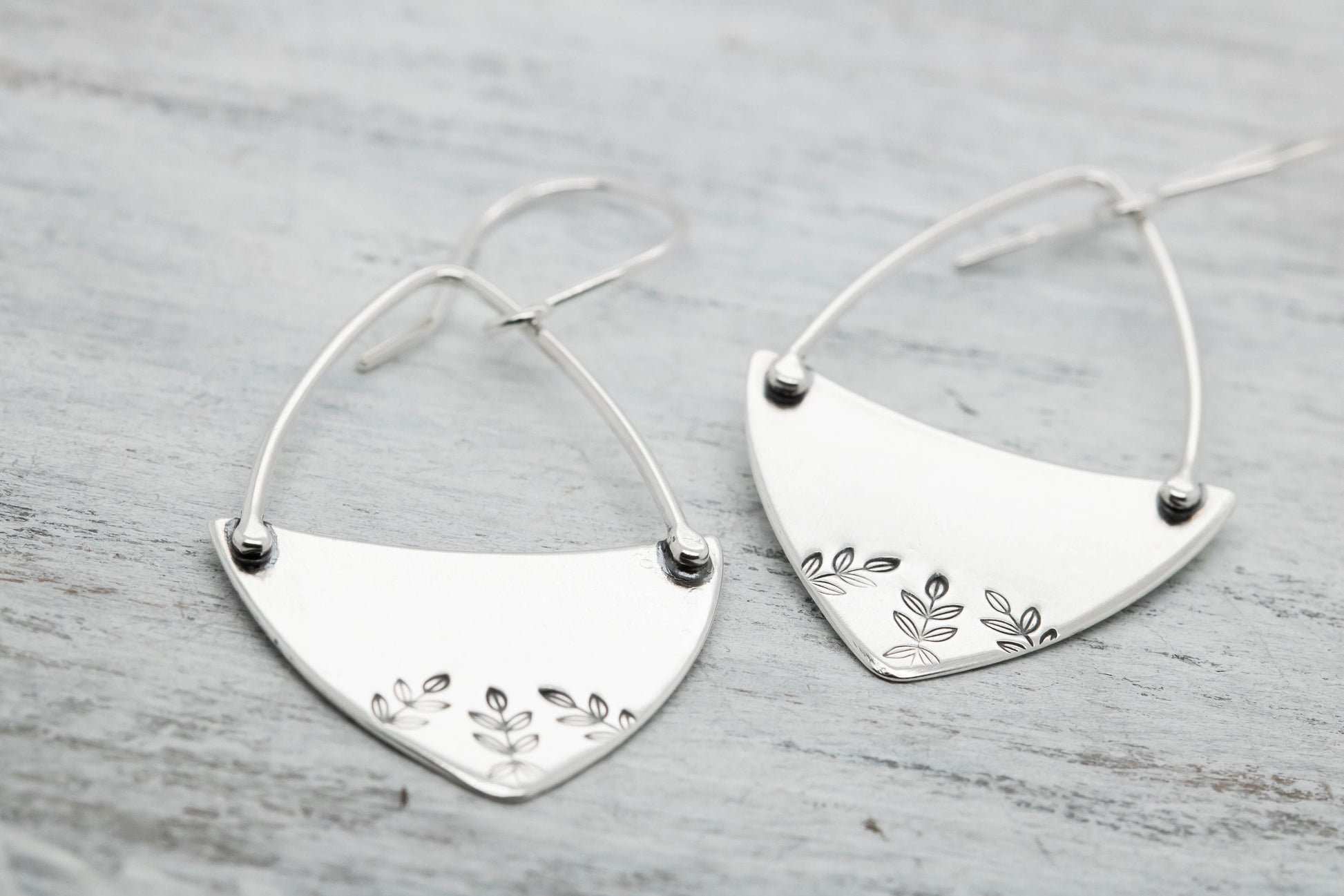 : Handcrafted sterling silver fern-stamped hoop earrings on a wooden surface, showcasing elegant leaf designs.
