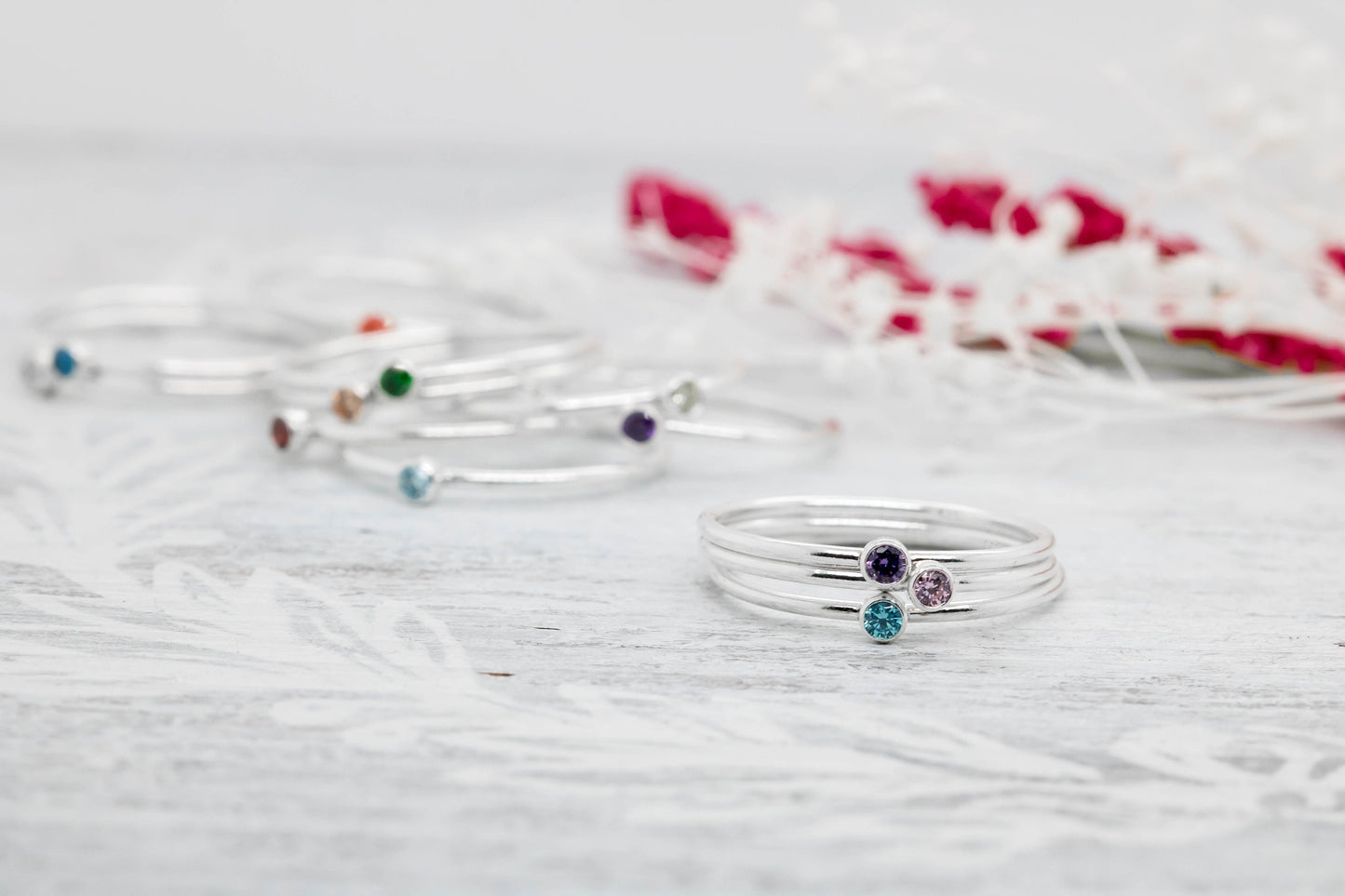 Three dainty sterling silver stacking rings with green, blue, orange, Purple and pink cubic zirconia birthstones, displayed on a white marble surface with pink flowers

