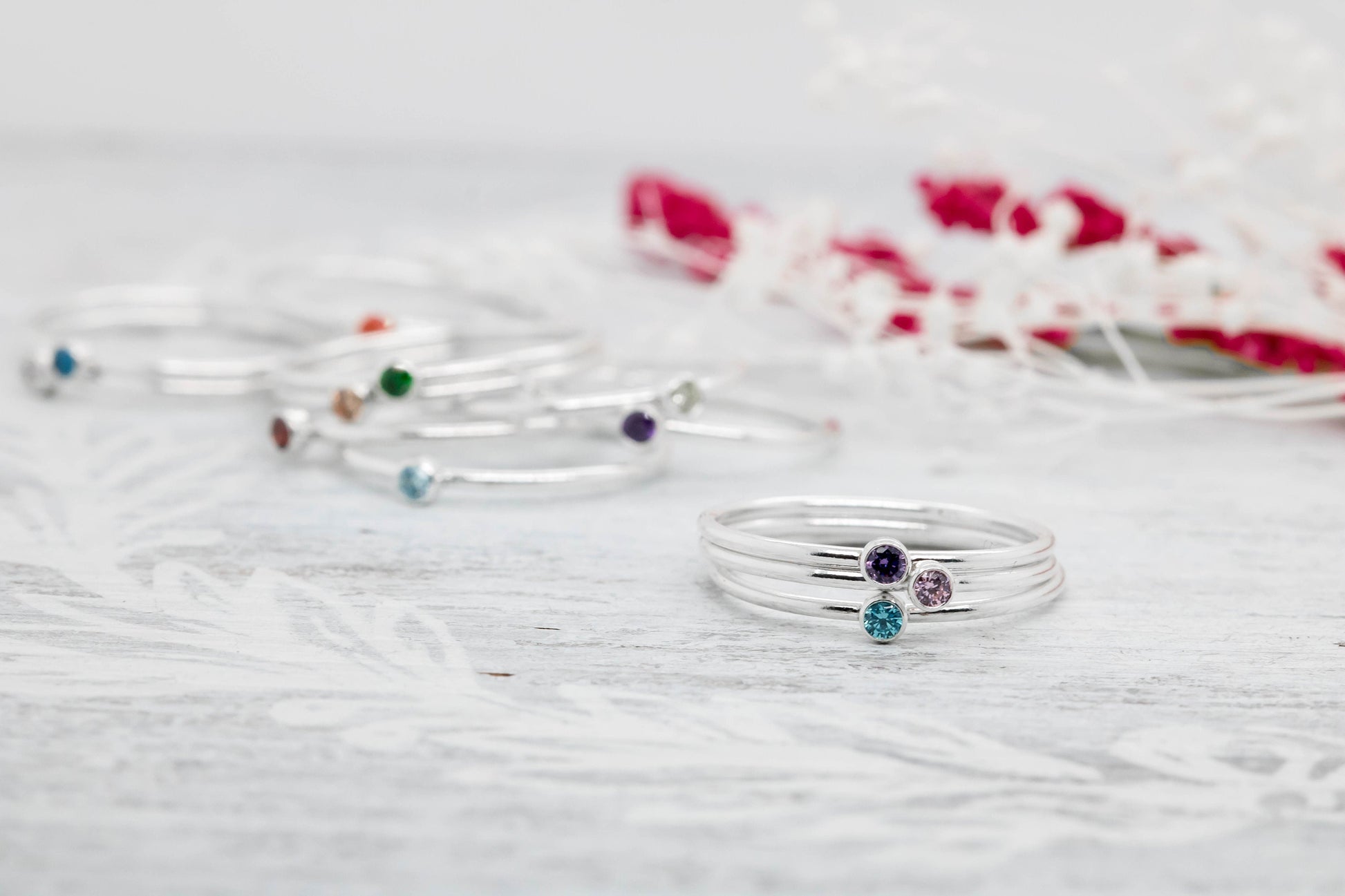 Three dainty sterling silver stacking rings with green, blue, orange, Purple and pink cubic zirconia birthstones, displayed on a white marble surface with pink flowers

