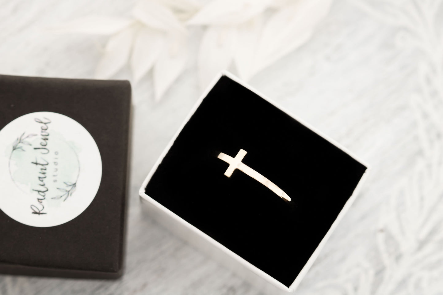 A solid 14k gold sideways cross ring displayed in an open black velvet box with a branded sticker, emphasizing its readiness as a gift on a white textured surface.