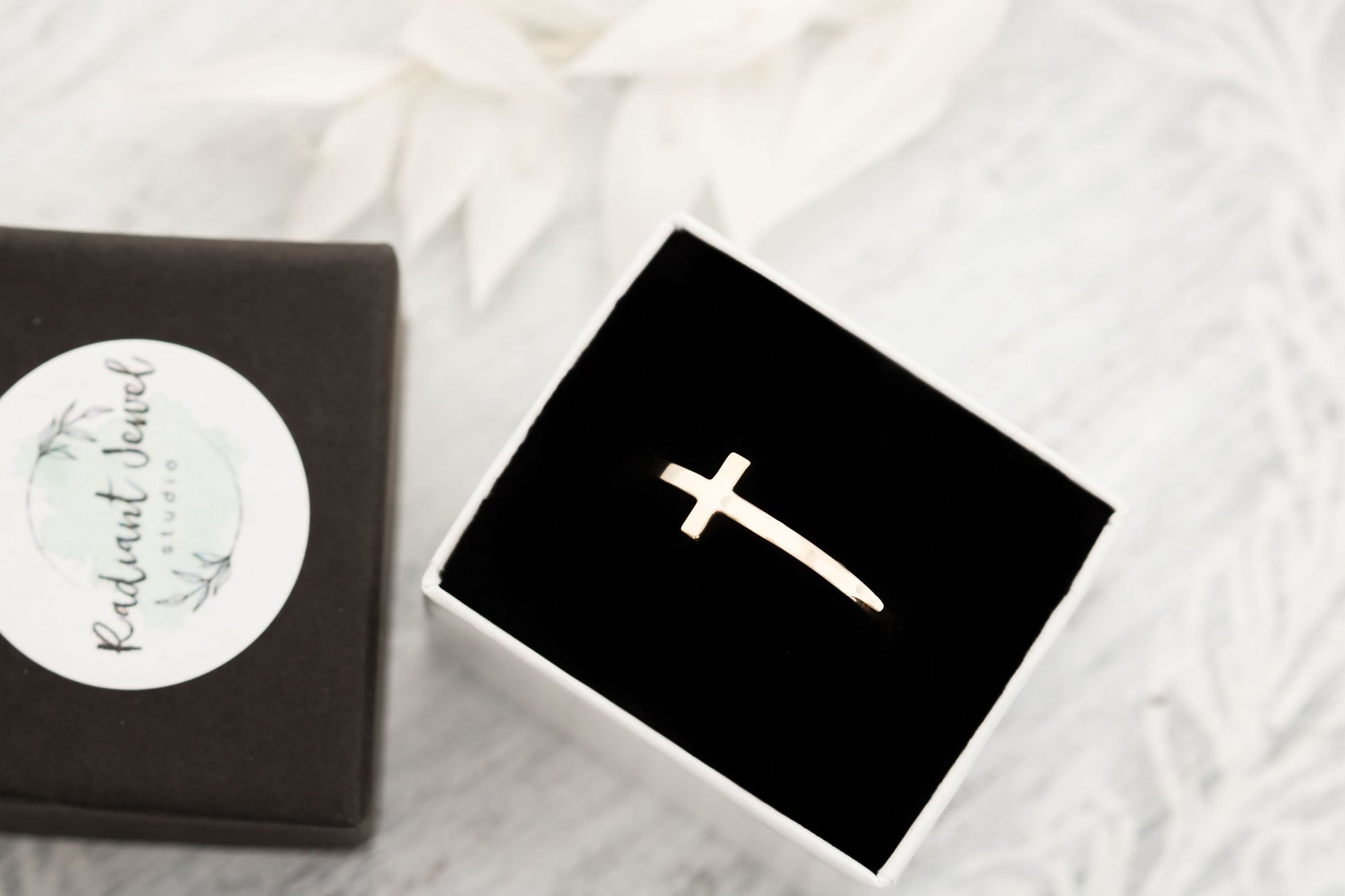 A solid 14k gold sideways cross ring displayed in an open black velvet box with a branded sticker, emphasizing its readiness as a gift on a white textured surface.