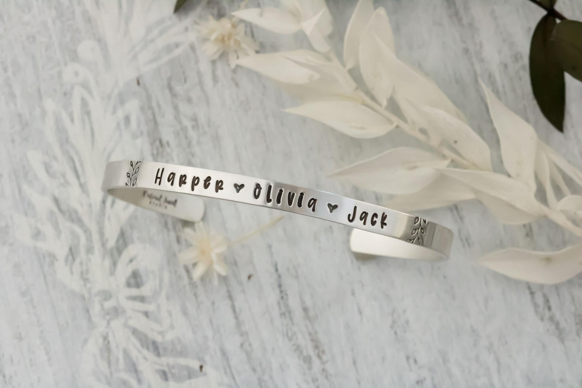 Sterling silver cuff bracelet stamped with kids names in a delicate handwritten font with a stamped for detail on both sides.